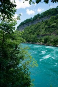 Turquoise river flows through lush forest, rocky cliff with foliage under partly cloudy sky.