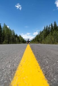 Person sitting on a road with yellow line, flanked by trees and mountains under blue sky.